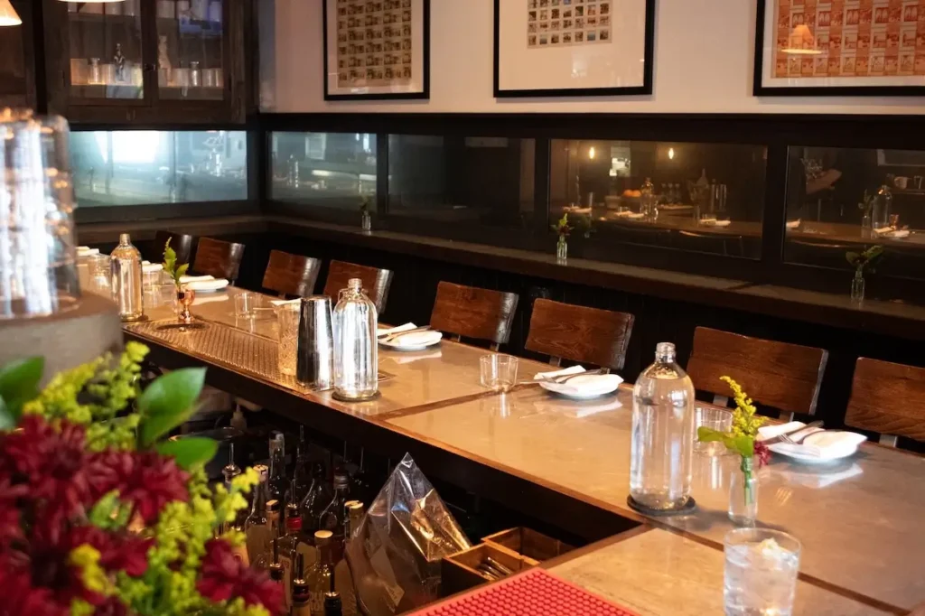 A polished restaurant or upscale bar interior with a long marble countertop set for dining. There are neatly arranged wooden chairs on one side and various glassware, metal cocktail shakers, and water carafes on the counter. Behind the bar, shelves hold bottles and tools, and the background wall features framed art and a window into a kitchen or prep area. A small flower arrangement and a glass of water sit in the foreground, while soft lighting creates a warm, inviting atmosphere.