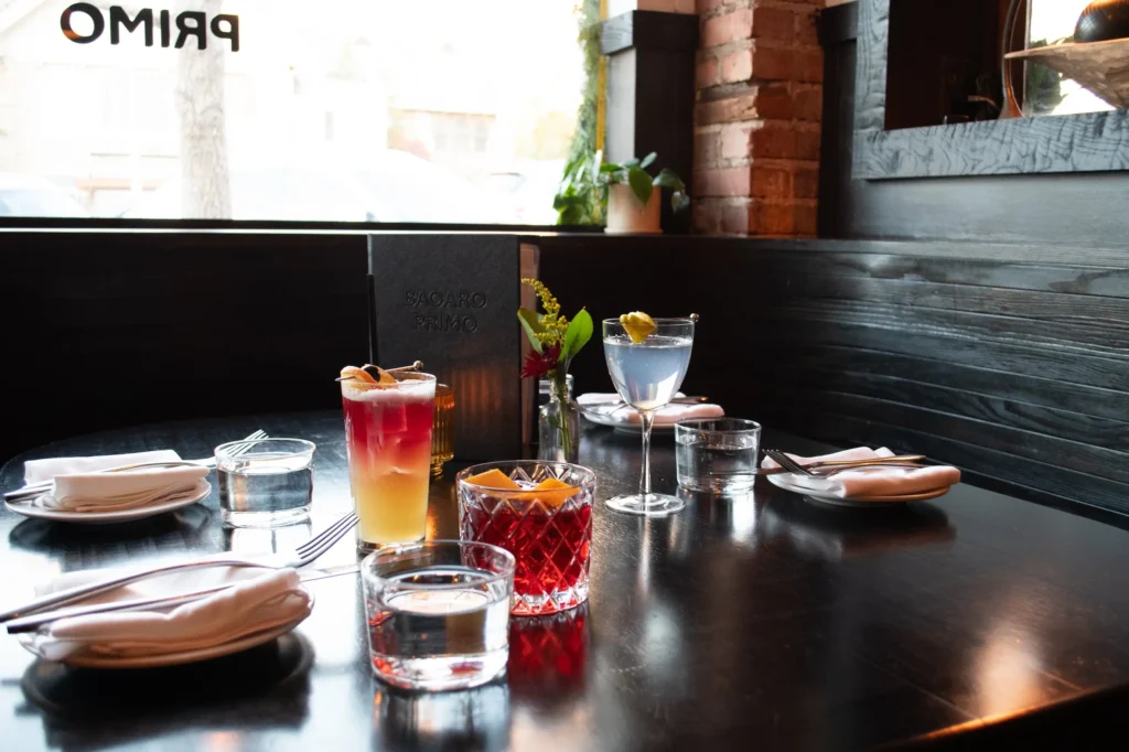 A dark wood table set for four in a cozy restaurant booth, featuring a variety of colorful cocktails and glass tumblers of water. The table is set with white napkins, forks, and small plates. Visible drinks include a layered yellow-to-red cocktail garnished with dried citrus in a tall glass, a purple martini with a lemon twist, and two cut-glass tumblers with red cocktails garnished with orange peel. A small vase with fresh flowers and a dark Bacaro Primo menu add decorative touches. The booth is against a window with 'PRIMO' written in reverse, and the background shows a brick wall, a mirror, and a potted plant on the window ledge.